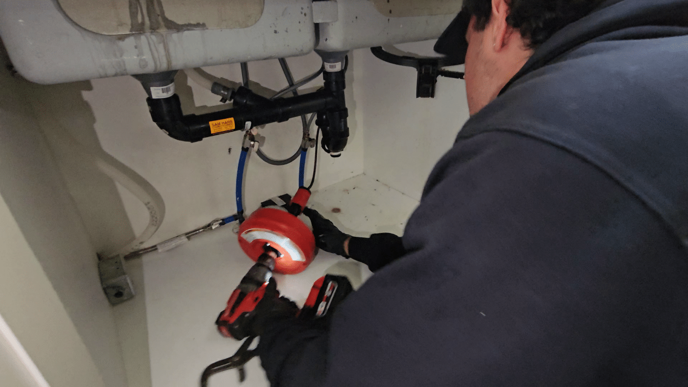 Environmental Plumbing technician using a powered drain snake beneath a kitchen sink
